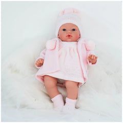 Koke doll in pink duffle coat and hat, lying on soft white fabric, showcasing realistic features and blue eyes.