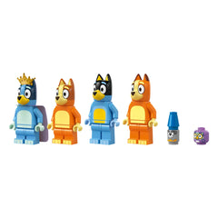 Four colorful LEGO minifigures from the Bluey series stand in a row: Bluey in blue with a crown, Bingo in orange, Bandit in black, and Chilli in orange. Additional accessories include a blue crayon and a purple block.