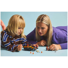 A child with blonde hair in a striped shirt and a woman with long blonde hair lean over a vibrant LEGO® City Wheel Loader set. They are playing on a soft blue surface, surrounded by construction accessories like traffic cones and a roadwork sign. The scene captures an engaging moment of imaginative play.