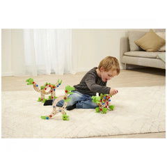 A child in blue jeans plays with a green and brown dinosaur construction set on a white rug indoors.