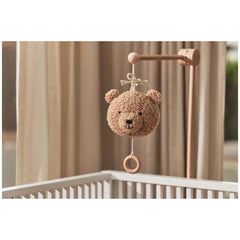 Brown teddy bear stuffed animal hanging from a brown wooden hook on a white railing with a beige curtain in the background.