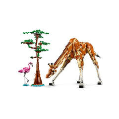 A posable LEGO giraffe with brown and white bricks leans towards a small brown tree and a pink flamingo on a blue base.