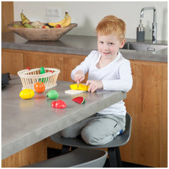 a bright kitchen with a smiling child in a white shirt and gray sweatpants, engaged with a colorful fruit bowl.