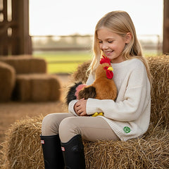 Plush rooster stuffed animal held by a girl in a white sweater and black boots, sitting on a brown hay bale.