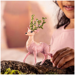 Pink Schleich Bayala Magic Deer figurine with green crown held by a child's hand against a blurred muted pink background.