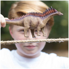 A child with blonde hair holds a textured brown Amargasaurus toy dinosaur in an outdoor setting.