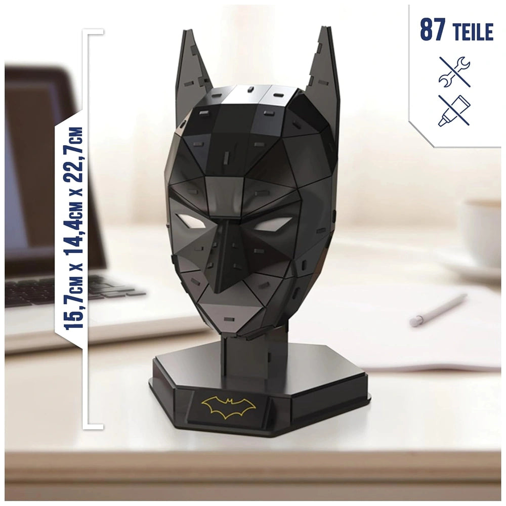 Batman mask 3D puzzle model kit on a black stand, featuring a textured design and set in a workspace with a laptop and cup.