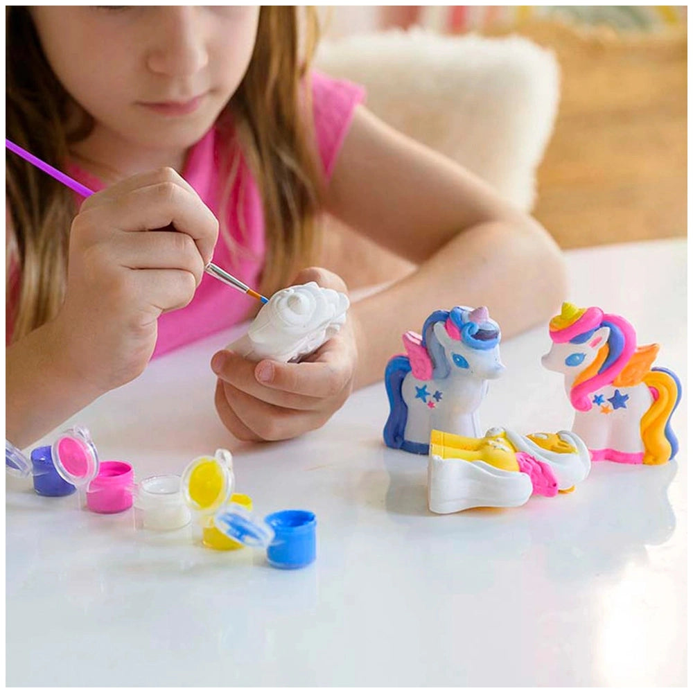 Young girl in pink top holding a glossy craft unicorn on a white tabletop with colorful craft items.