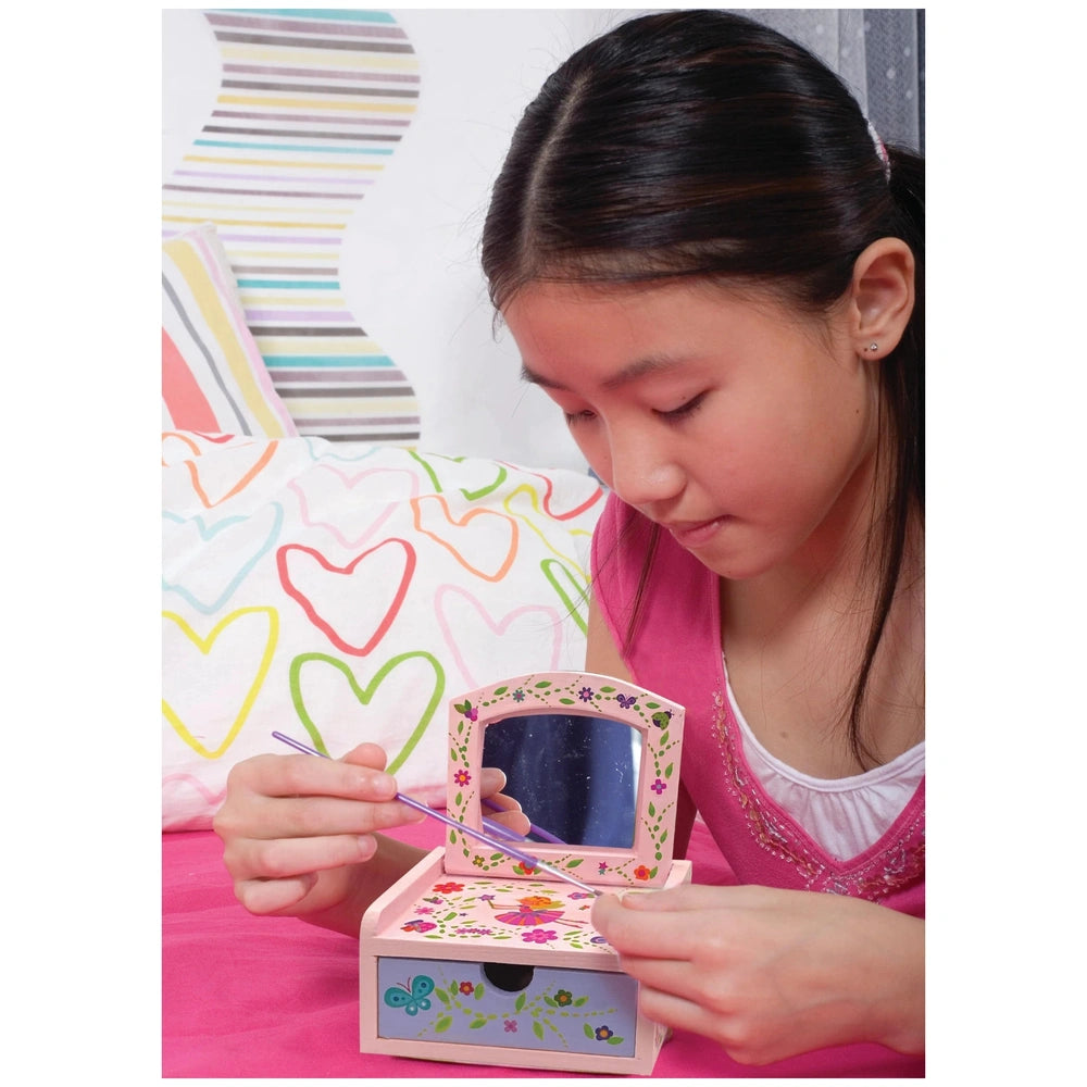 Girl in pink top holds a small pink fairy chest toy with a mirror, set against a colorful hearts patterned bedspread.