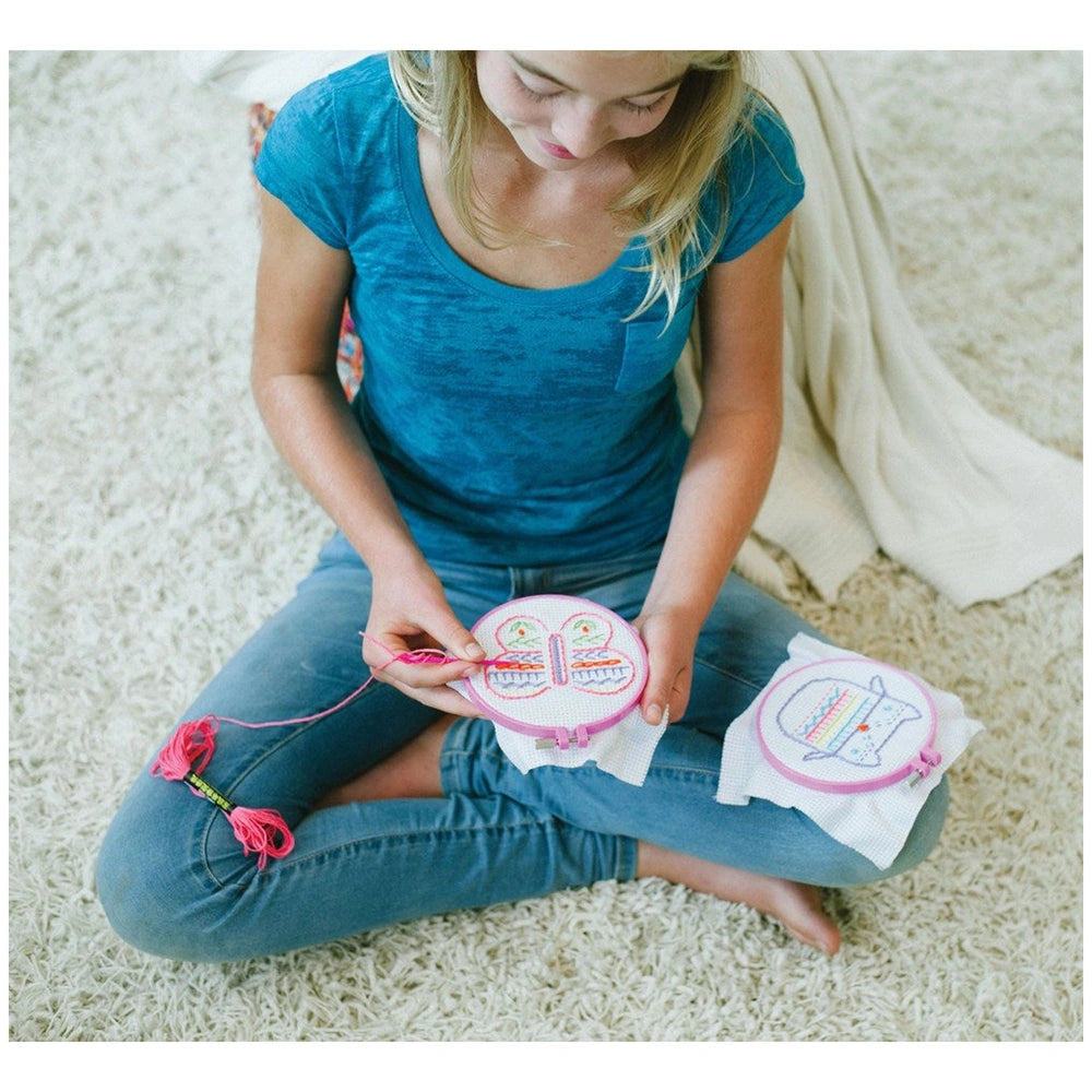 4M Embroidery Stitches kit with a woman in a blue t-shirt sitting on a white carpet, holding a pink embroidery hoop.