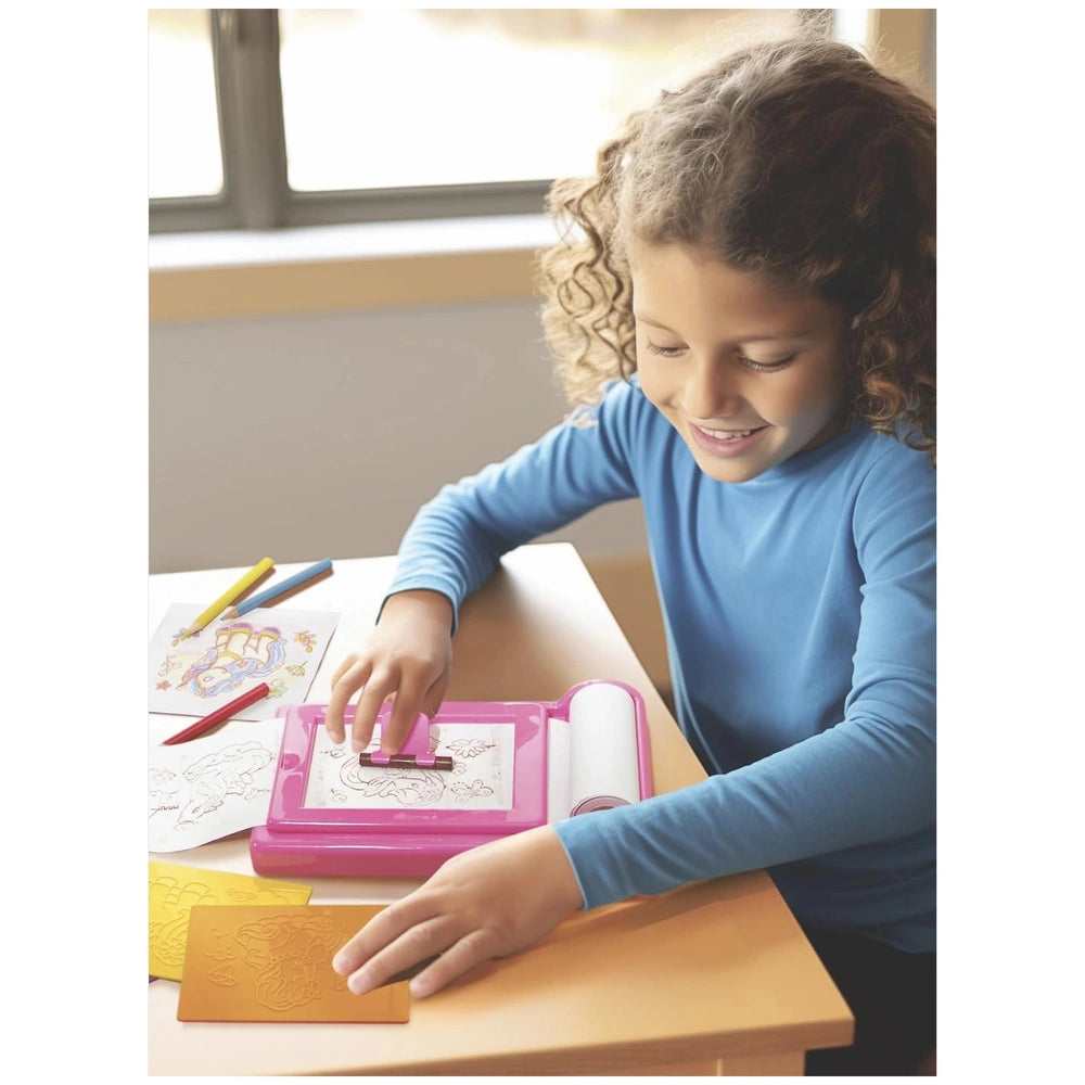 KidzMaker Unicorn Rub Art Studio with a young girl in a blue top using a pink drawing tool on a light wood tabletop.