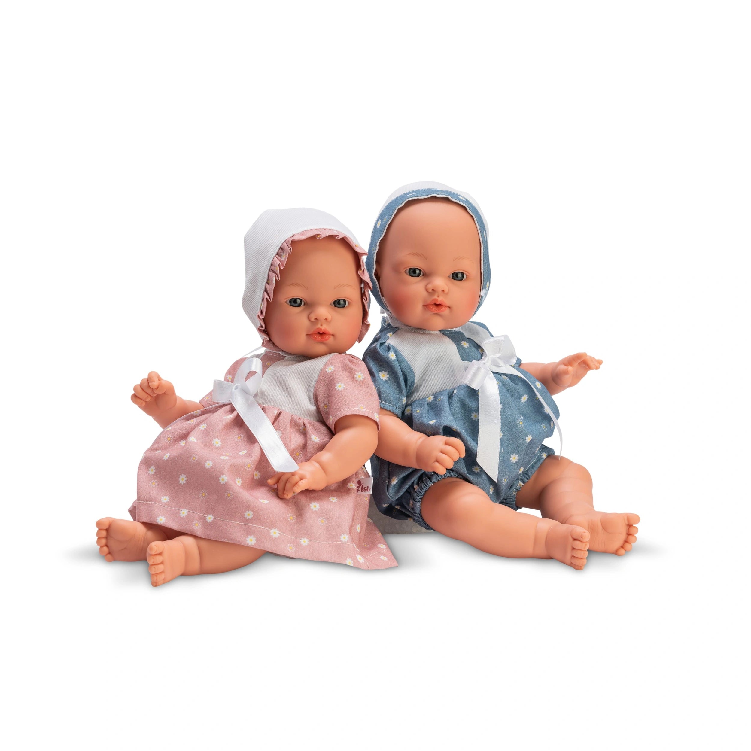 Two ASI Dolls in daisy dresses; one pink and one blue, with matching bonnets and delicate features, sitting together.