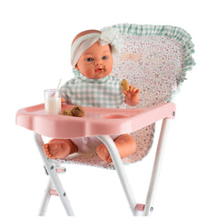 Doll in pink high chair with cookies and milk, wearing a gingham bib and headband with floral background.