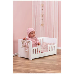 Doll in a white wooden cradle with floral bedding, set in a pink-themed room with sheer curtains.