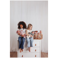 Two children sitting on a white dresser, each holding a doll from ASI's Iris Collection, with a basket of dolls beside them.