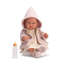 ASI Dolls María, 43 cm, in a soft pink coat and hood, white dress and booties, sits next to a baby bottle.