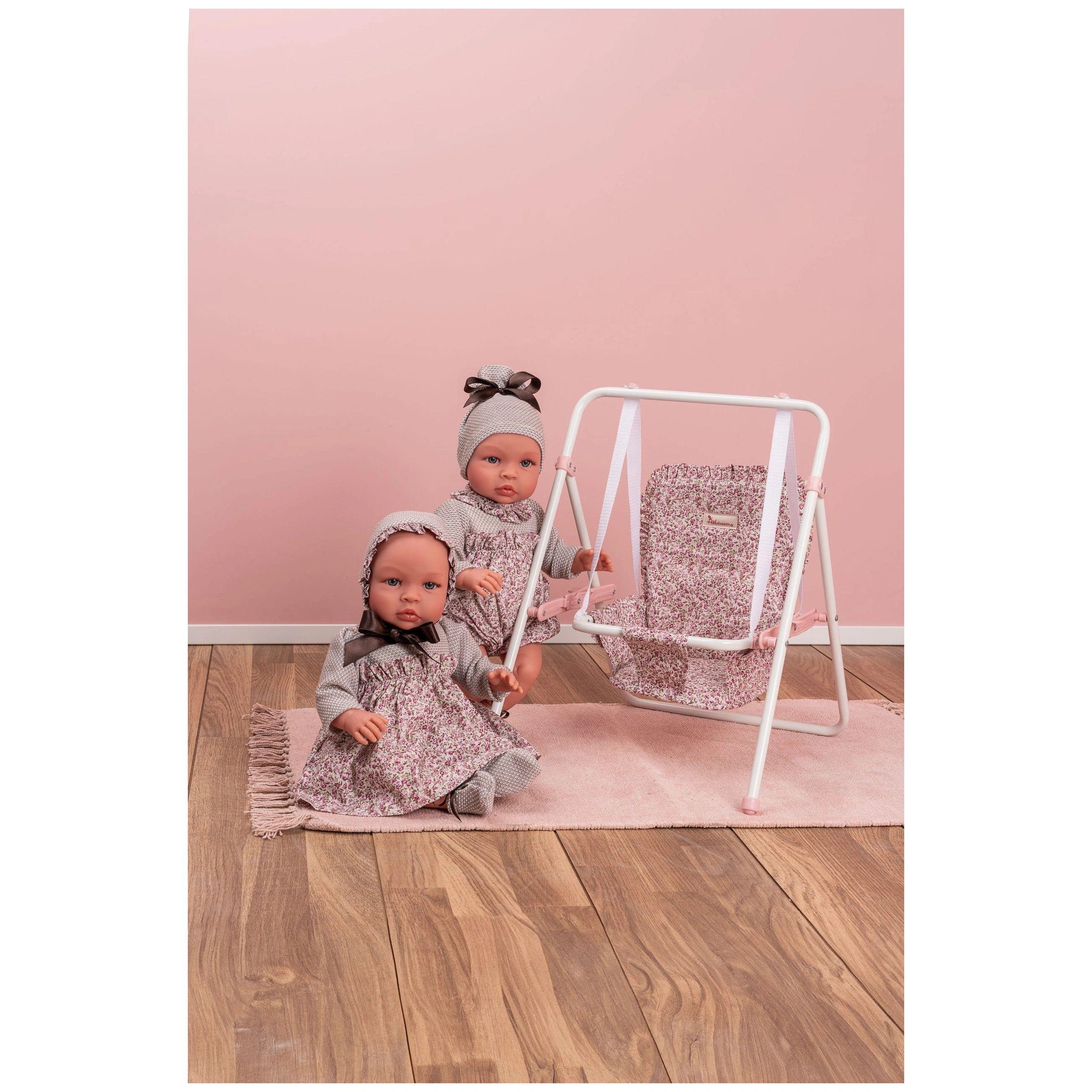 Two dolls in floral outfits sit next to a matching pink and white doll swing on a wooden floor.