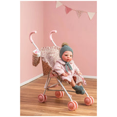 Doll in pink floral stroller with safety straps, pink wheels, and matching bag against a pastel pink wall with bunting.