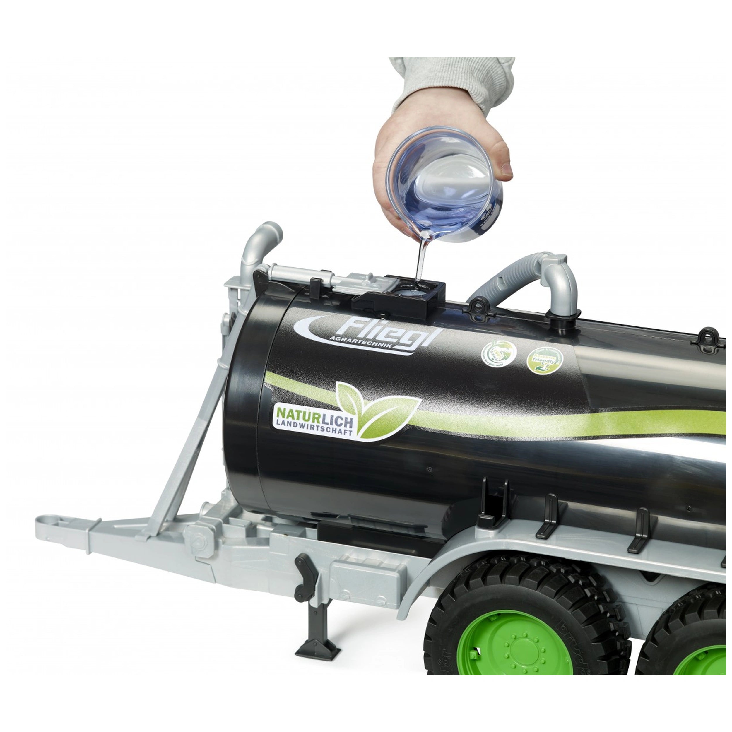 Hand pouring water into the black Bruder Fliegl Barrel Trailer Jumbo Line with green wheels, simulating farming play.