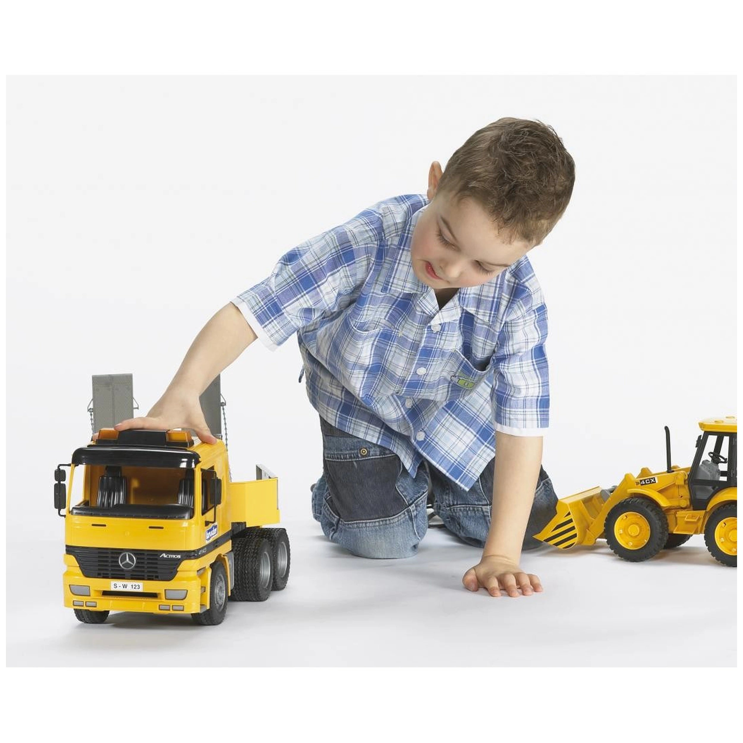 Child in plaid shirt playing with yellow Bruder JCB 4CX backhoe loader and toy truck on a white background.