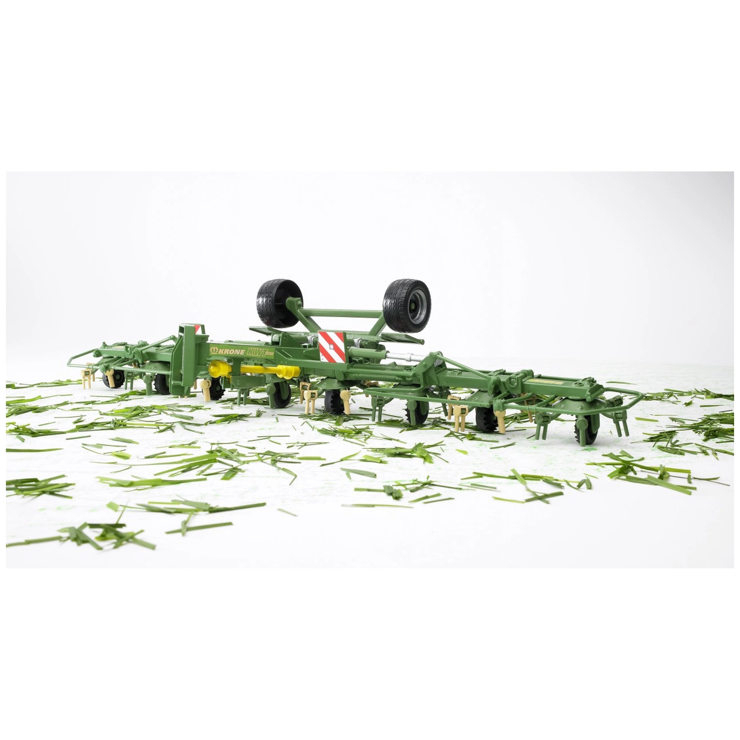 Bruder Krone Trailed Rotary Tedder toy, green with wheels, on white surface scattered with grass.
