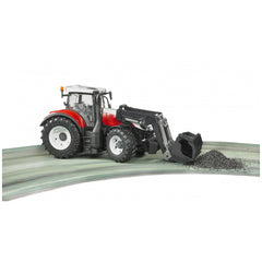 Red and black Bruder Steyr 6300 Terrus CVT toy tractor with front loader moving gravel on a curved road.