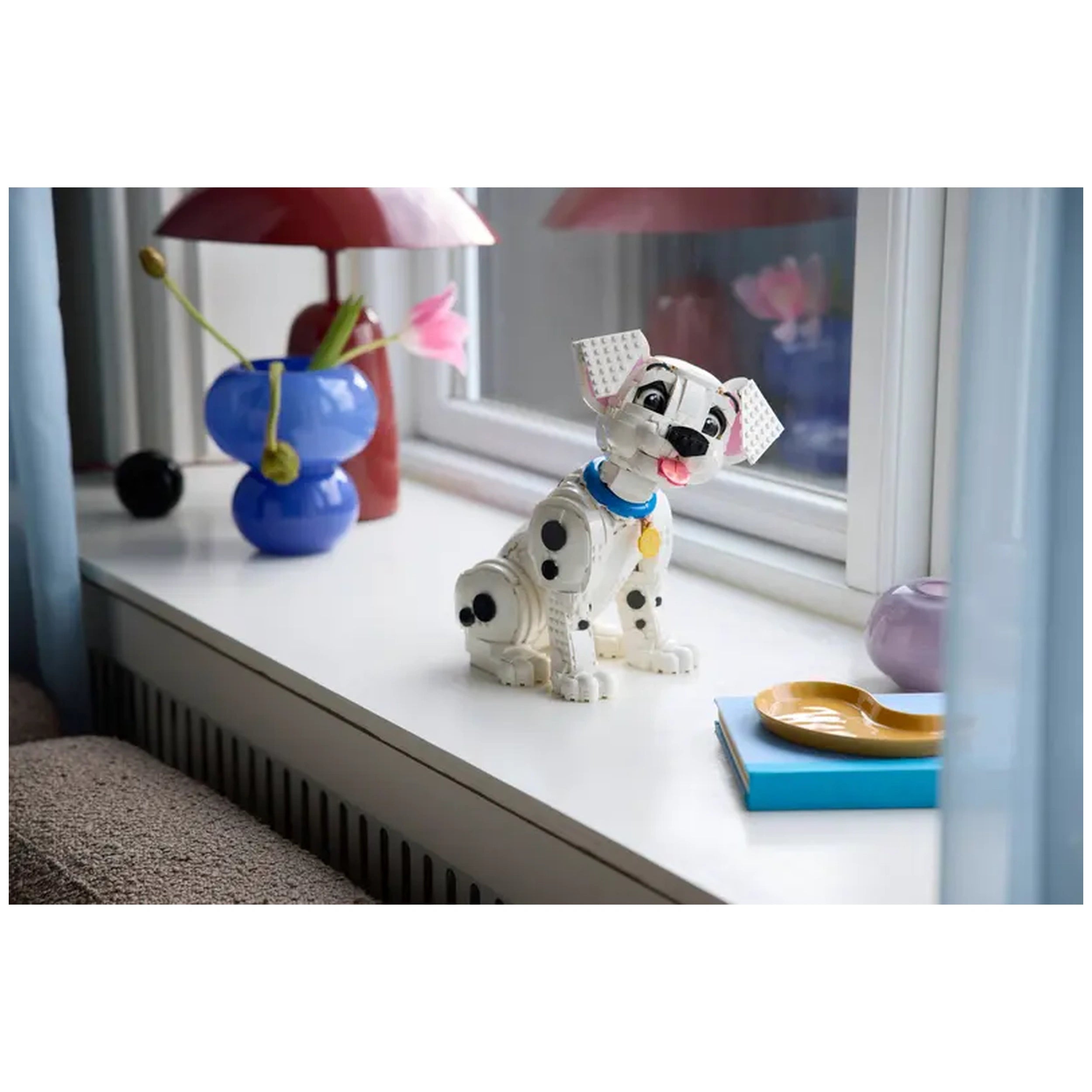 A playful Dalmatian puppy LEGO model sits on a white windowsill, next to a colorful vase with flowers and a bright lamp.