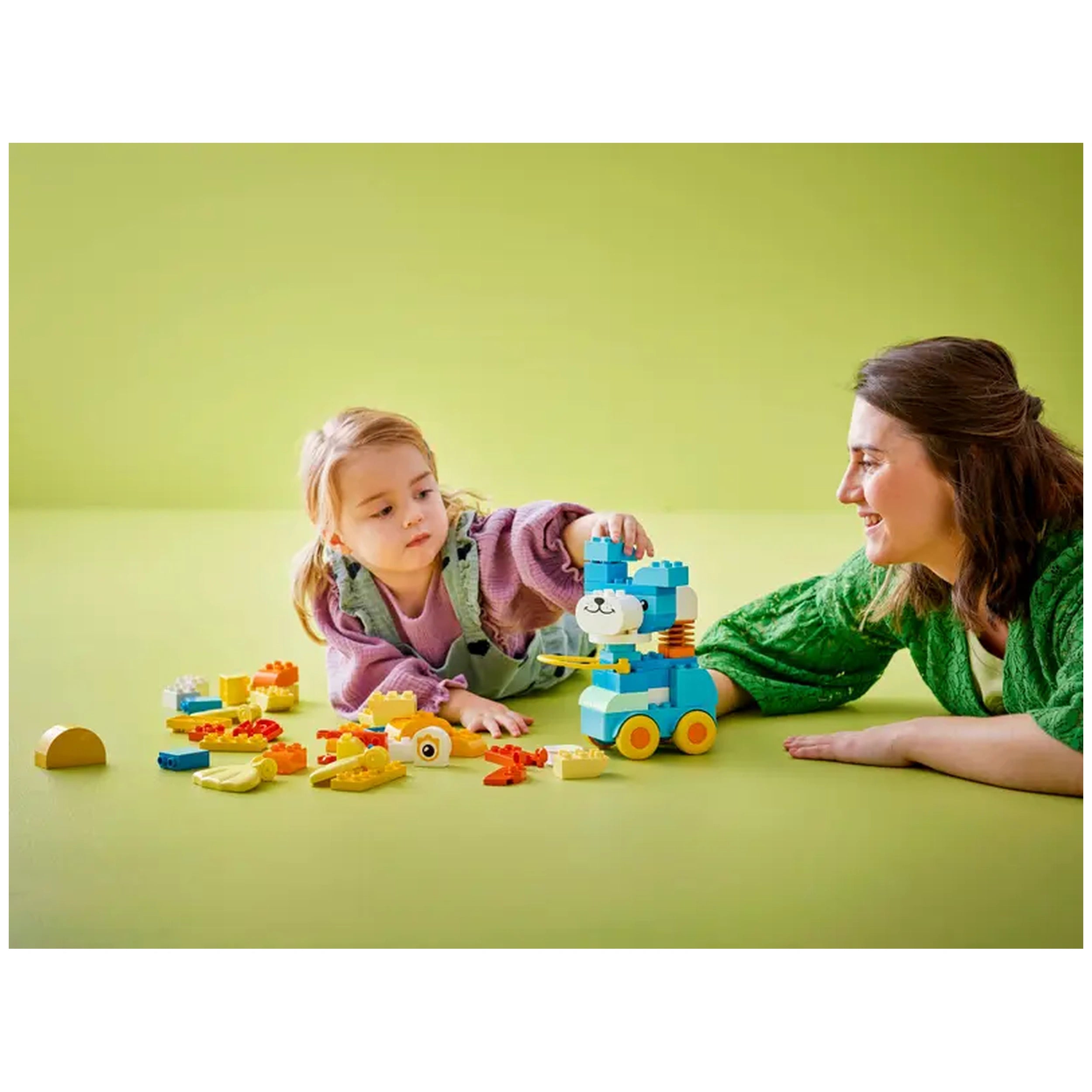 A young girl in a lavender sweater builds a colorful LEGO animal on wheels with a smiling woman beside her. Various LEGO pieces are scattered on a soft green surface.