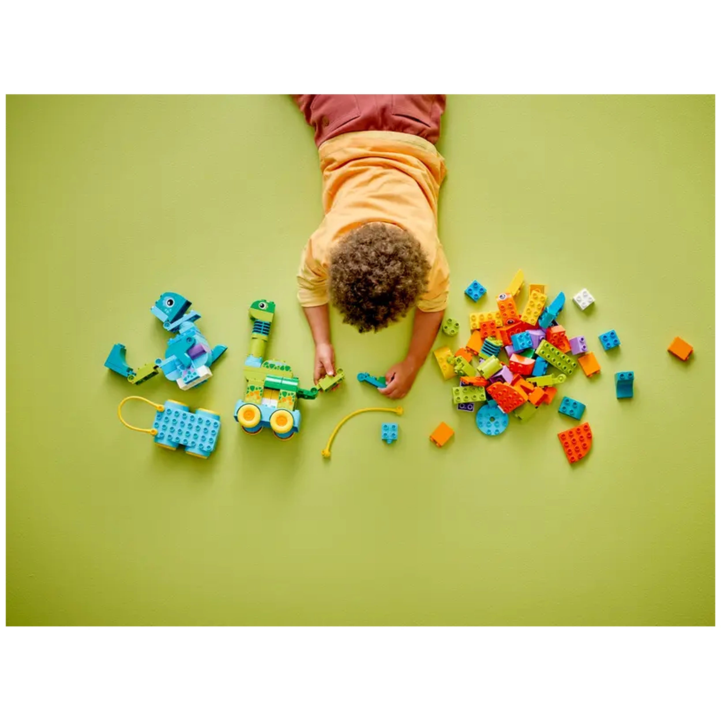 A child in a yellow shirt plays on a green surface, building with colorful LEGO bricks. The scene features a constructed dinosaur vehicle alongside scattered blocks, showcasing creativity and engagement.