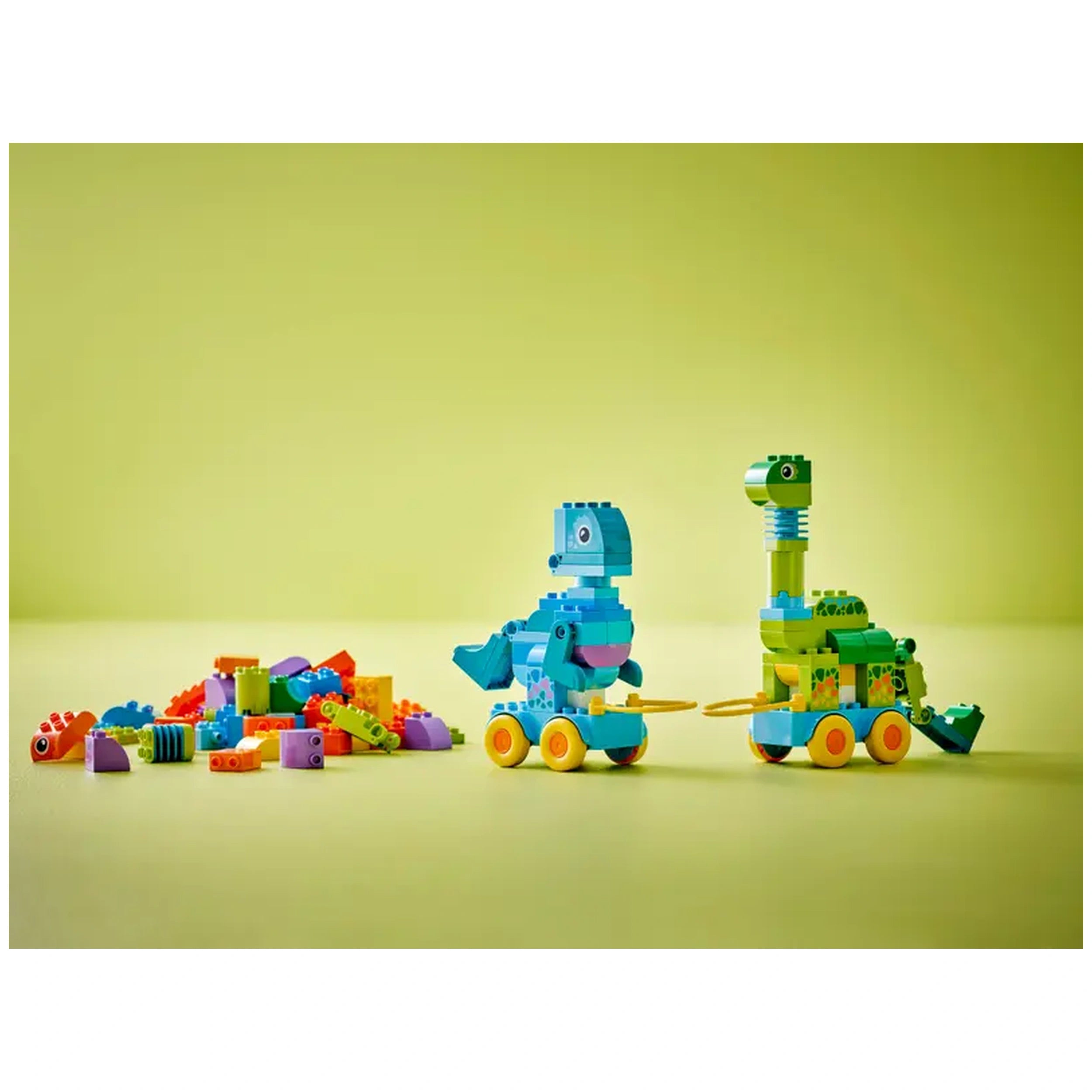 Colorful LEGO DUPLO dinosaur models on wheels, including a blue dinosaur and a green dinosaur, beside a pile of assorted bricks on a light green background.