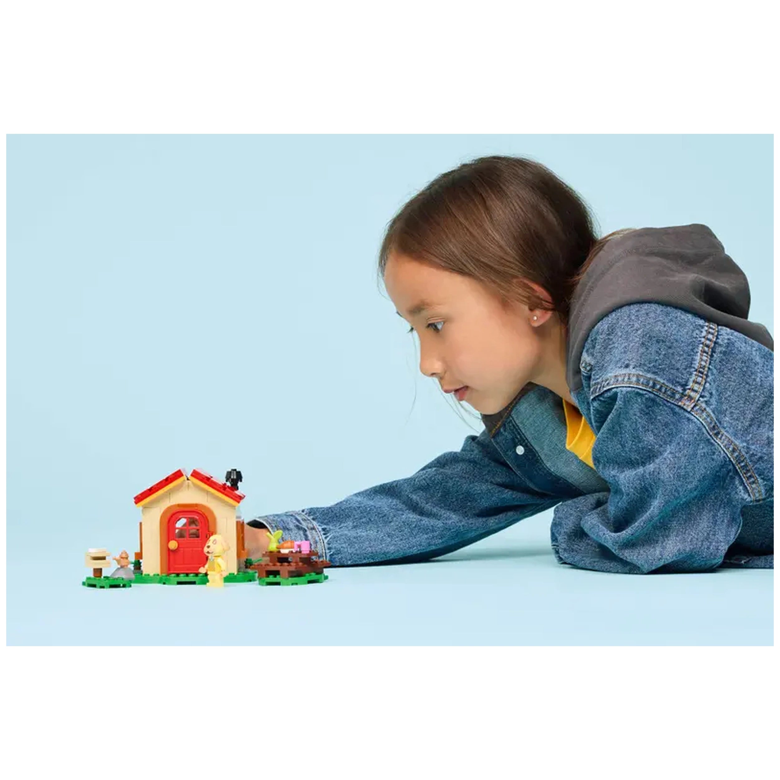 A child interacts with the LEGO - Goldie's Cozy House set, featuring a colorful building with a red door and vibrant outdoor elements like a picnic table and minifigures. The child is wearing a denim jacket and appears engaged in imaginative play, set against a light blue background.