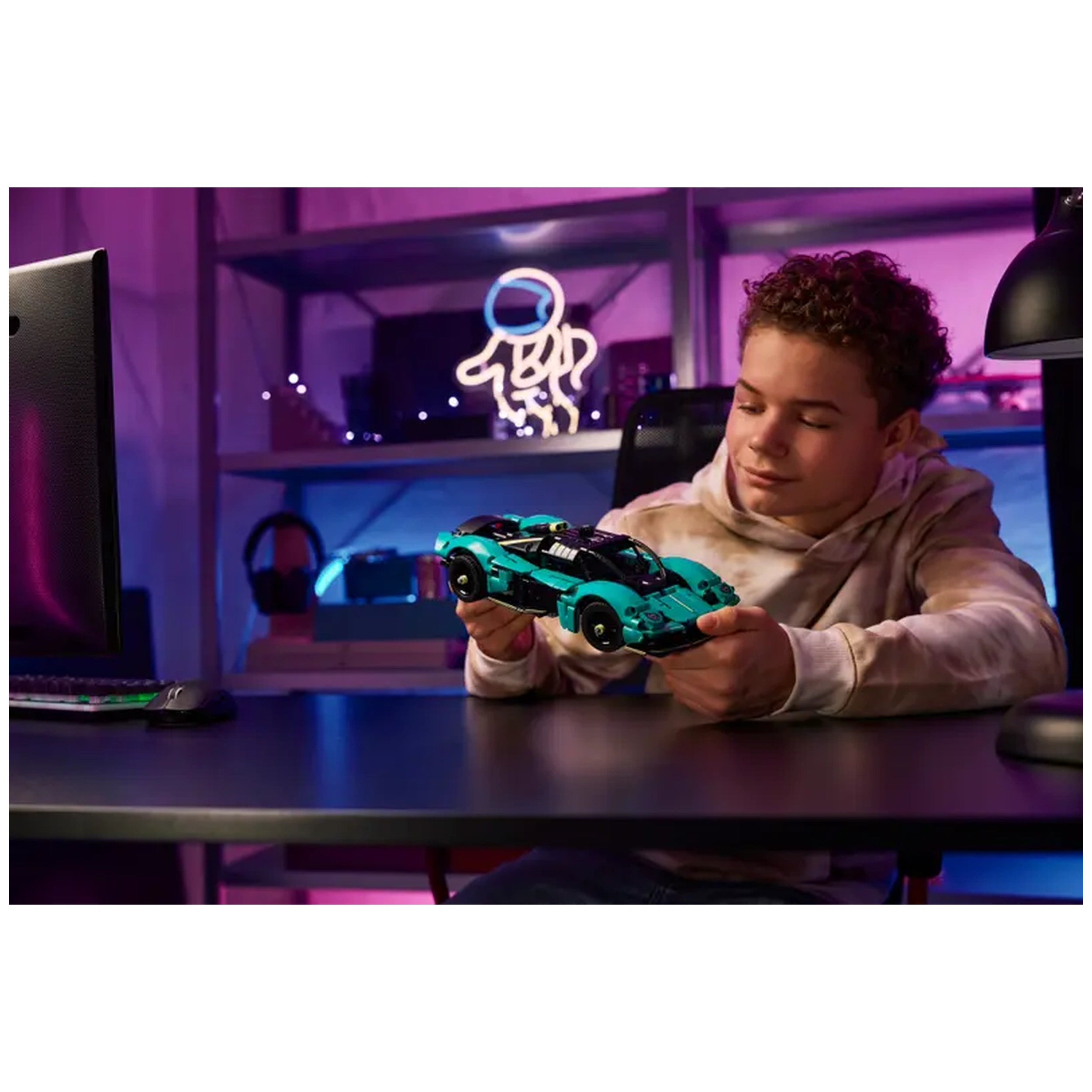 A boy sits at a desk, admiring the vibrant LEGO Aston Martin Valkyrie construction set in his hands, with colorful neon lights in the background.