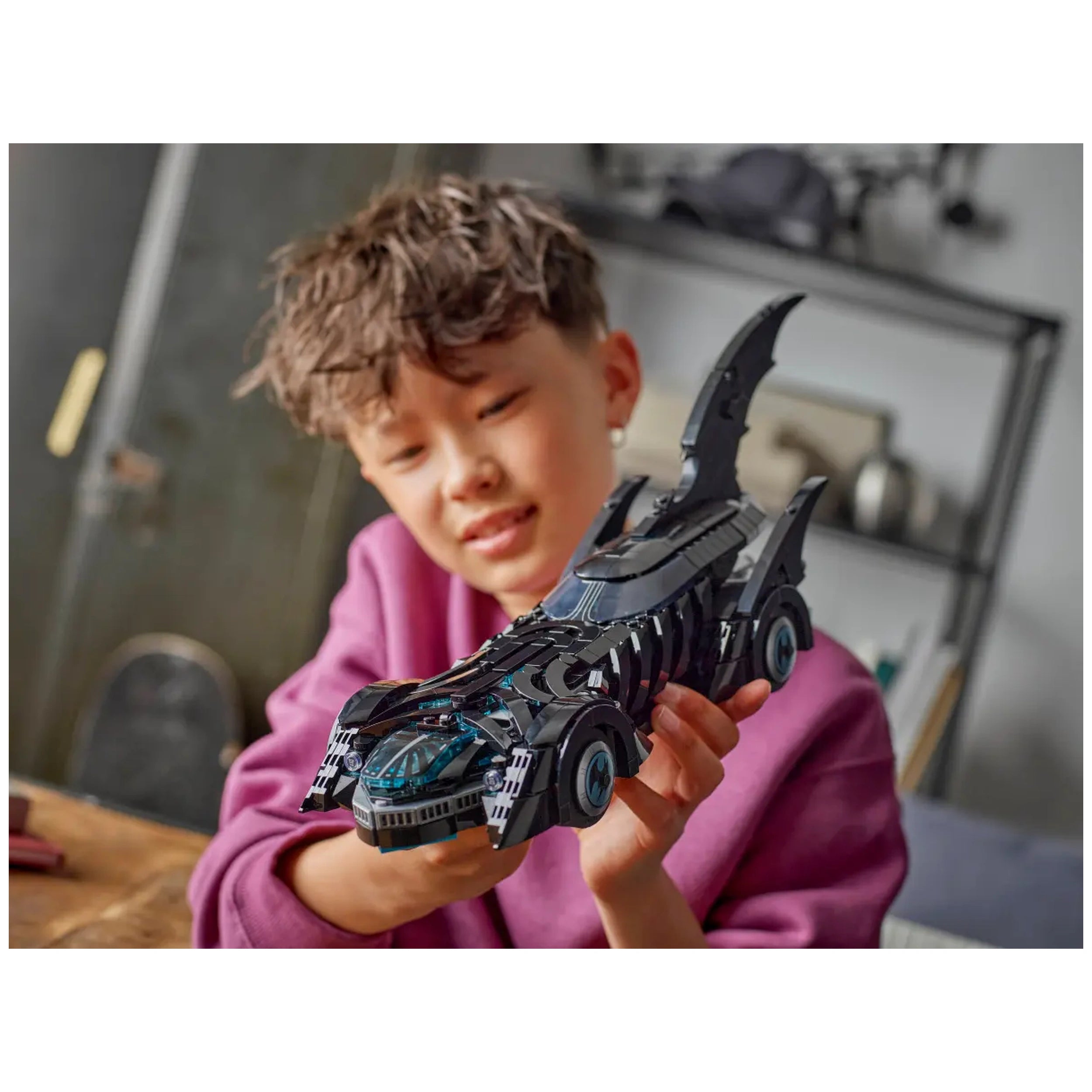 A child with curly hair smiles while holding the black LEGO® Batman Forever™ Batmobile™, showcasing its detailed design and blue accents.