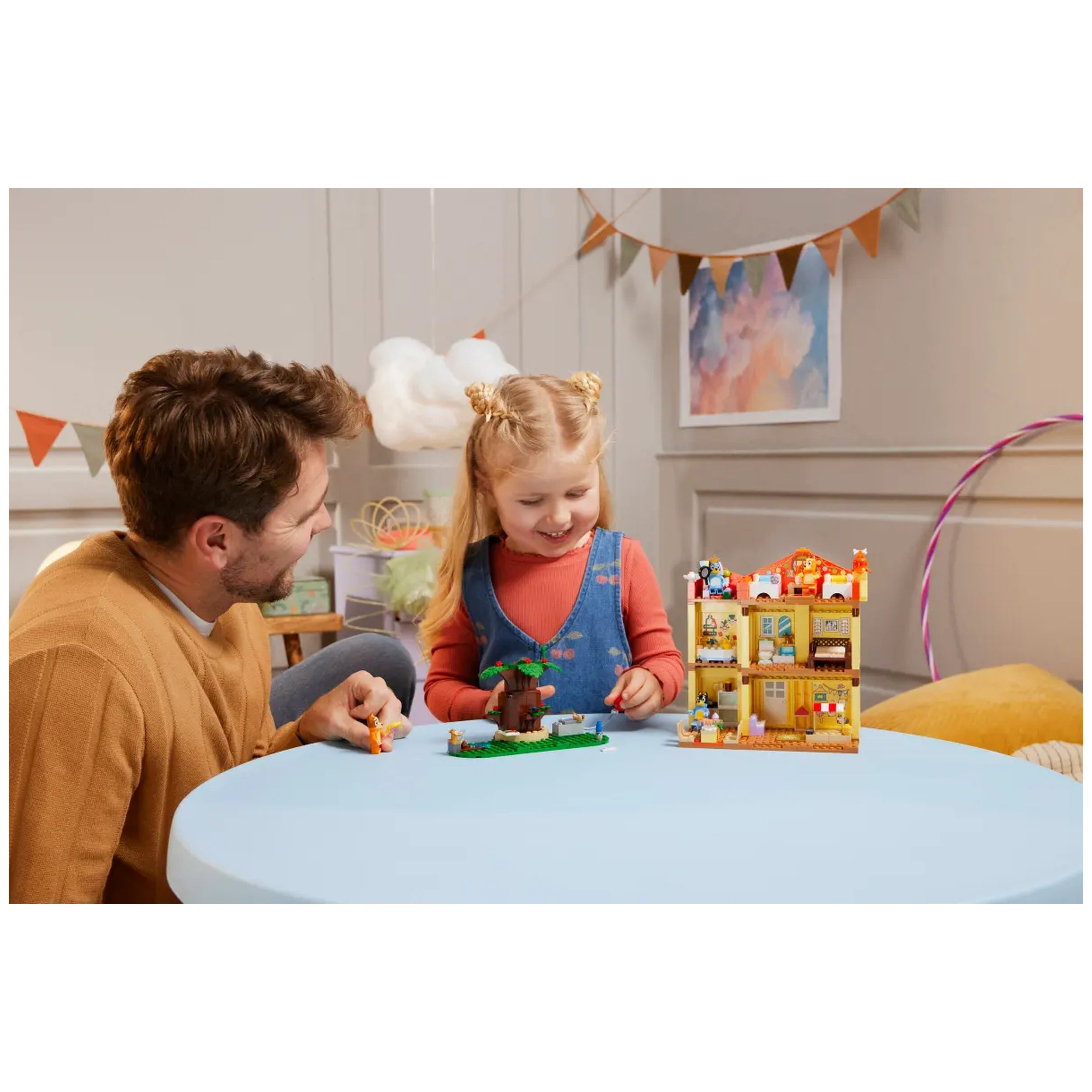 A child and a man enjoy building the LEGO Bluey’s Family House playset at a light blue table. The playset features a colorful house and various accessories, with the child smiling and interacting with the figures and elements. A playful and creative atmosphere is enhanced by colorful decor in the background.