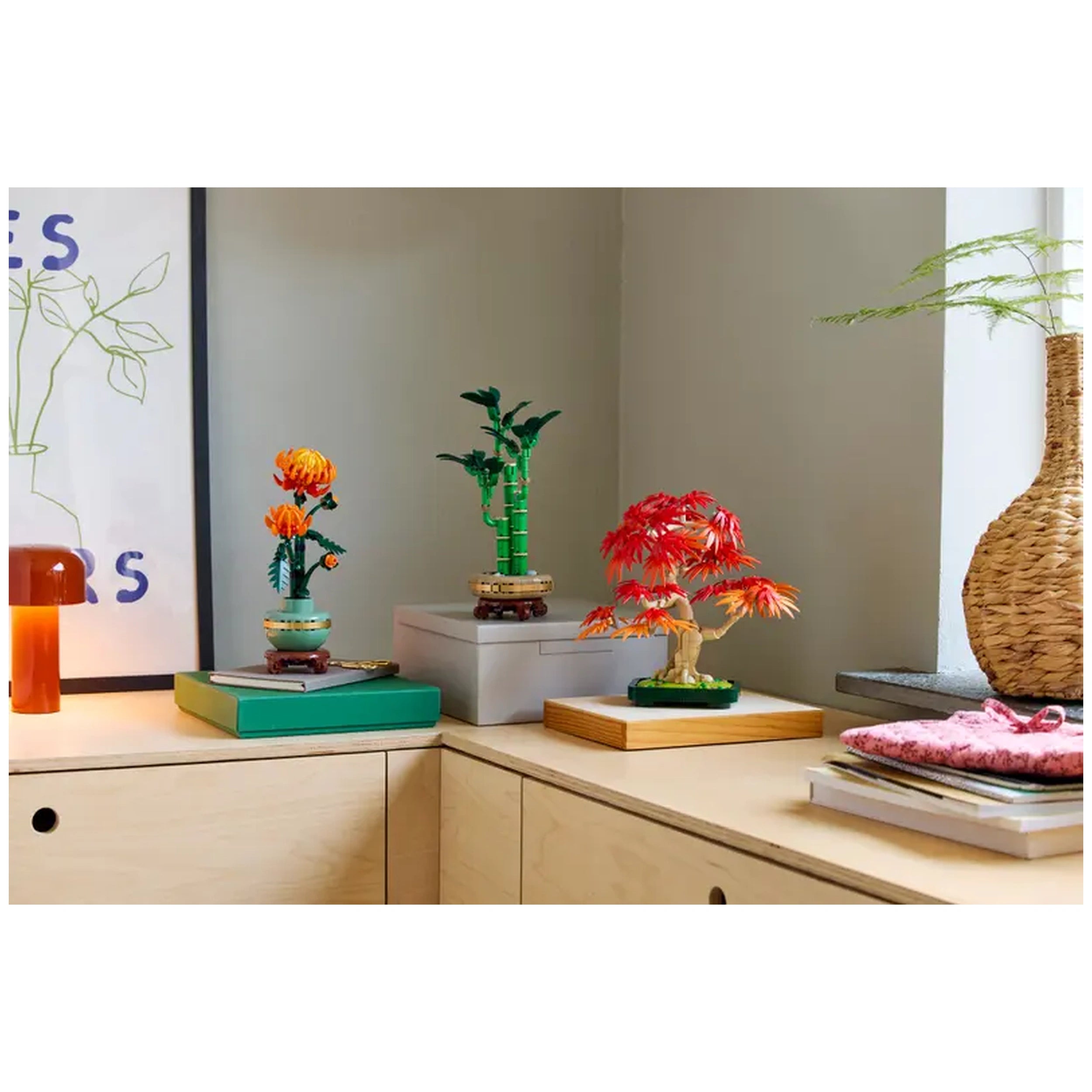Three vibrant LEGO® Botanicals sets displayed on a light wooden table. On the left, a colorful chrysanthemum in a pot; center, a green bamboo plant; and on the right, a striking Japanese Red Maple bonsai tree with red and orange leaves. A cozy ambiance enhanced by a warm-toned lamp and a woven vase in the background.
