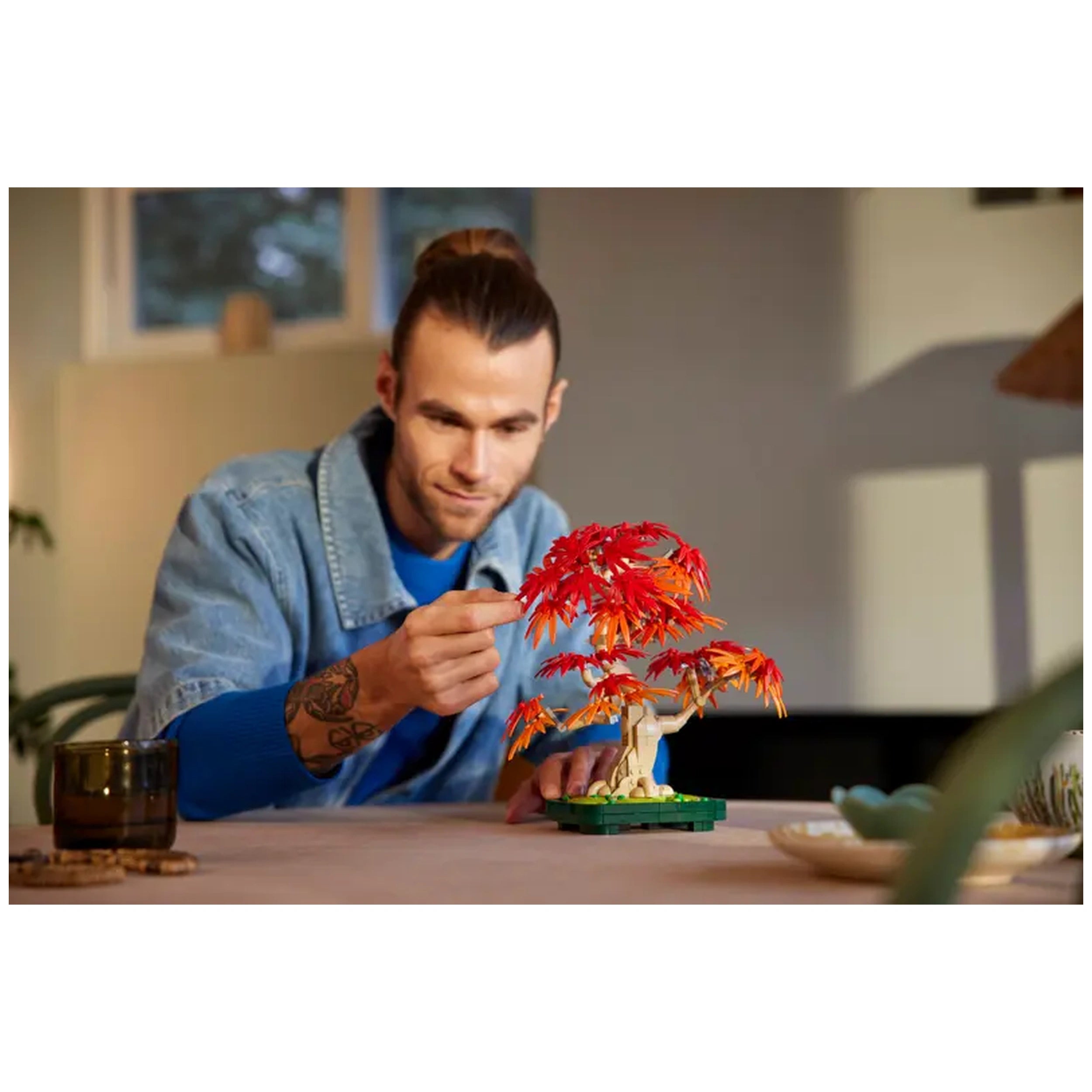 A man inspects the vibrant red and orange leaves of a LEGO Bonsai Tree on a tabletop, with a cozy interior in the background.