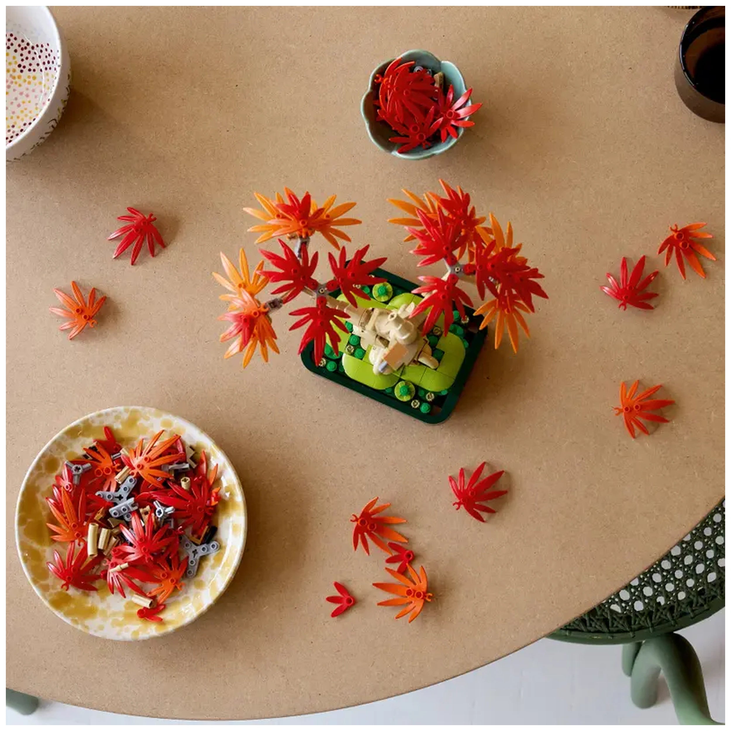 LEGO Botanicals Japanese Red Maple Bonsai Tree set on a light brown table, surrounded by vibrant red and orange leaves and building pieces. A decorative bowl holds additional leaf pieces, showcasing the intricate design elements of this creative construction set.