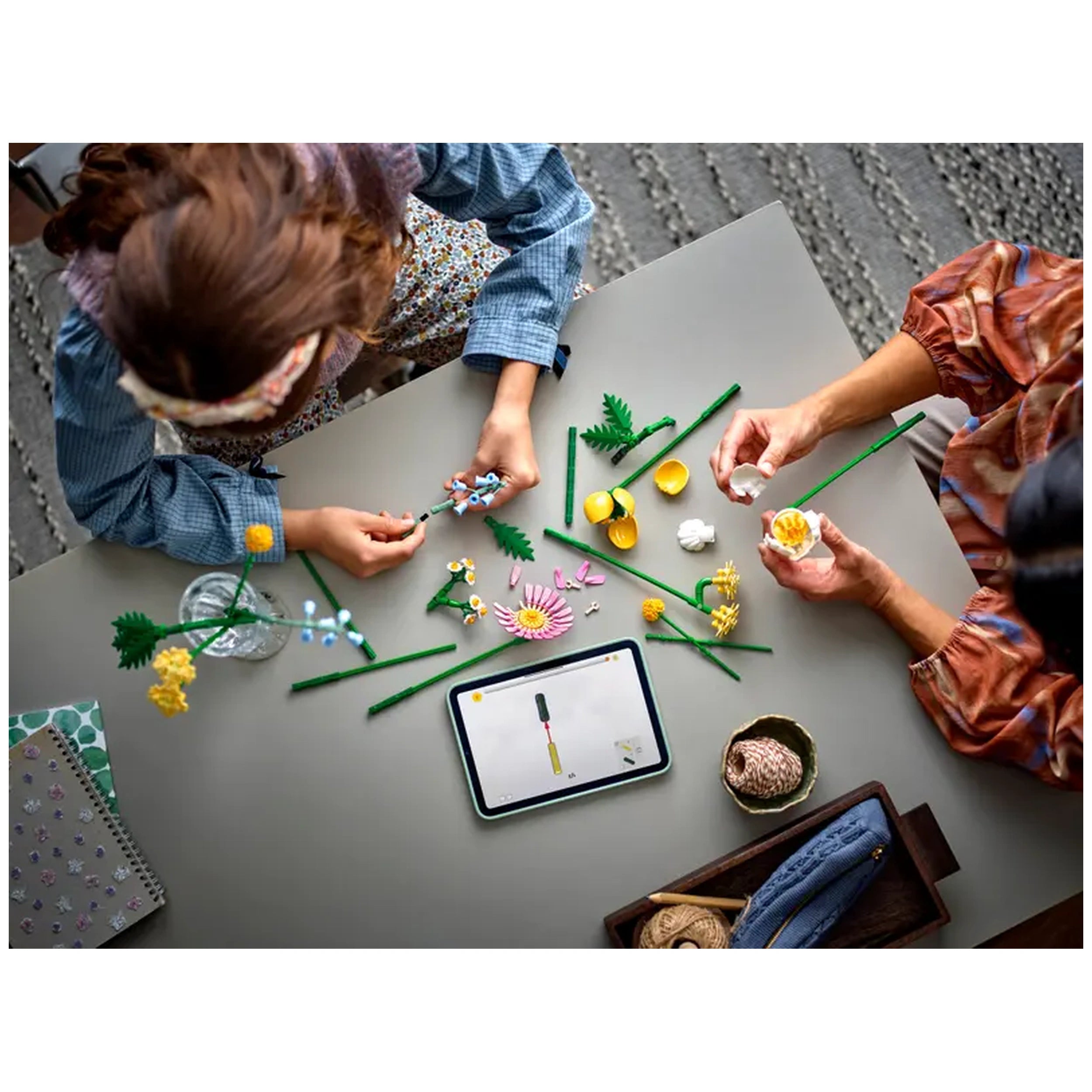 Two people assembling the LEGO Botanicals Petite Sunny Bouquet on a grey table, surrounded by colorful flower pieces and a tablet displaying instructions.