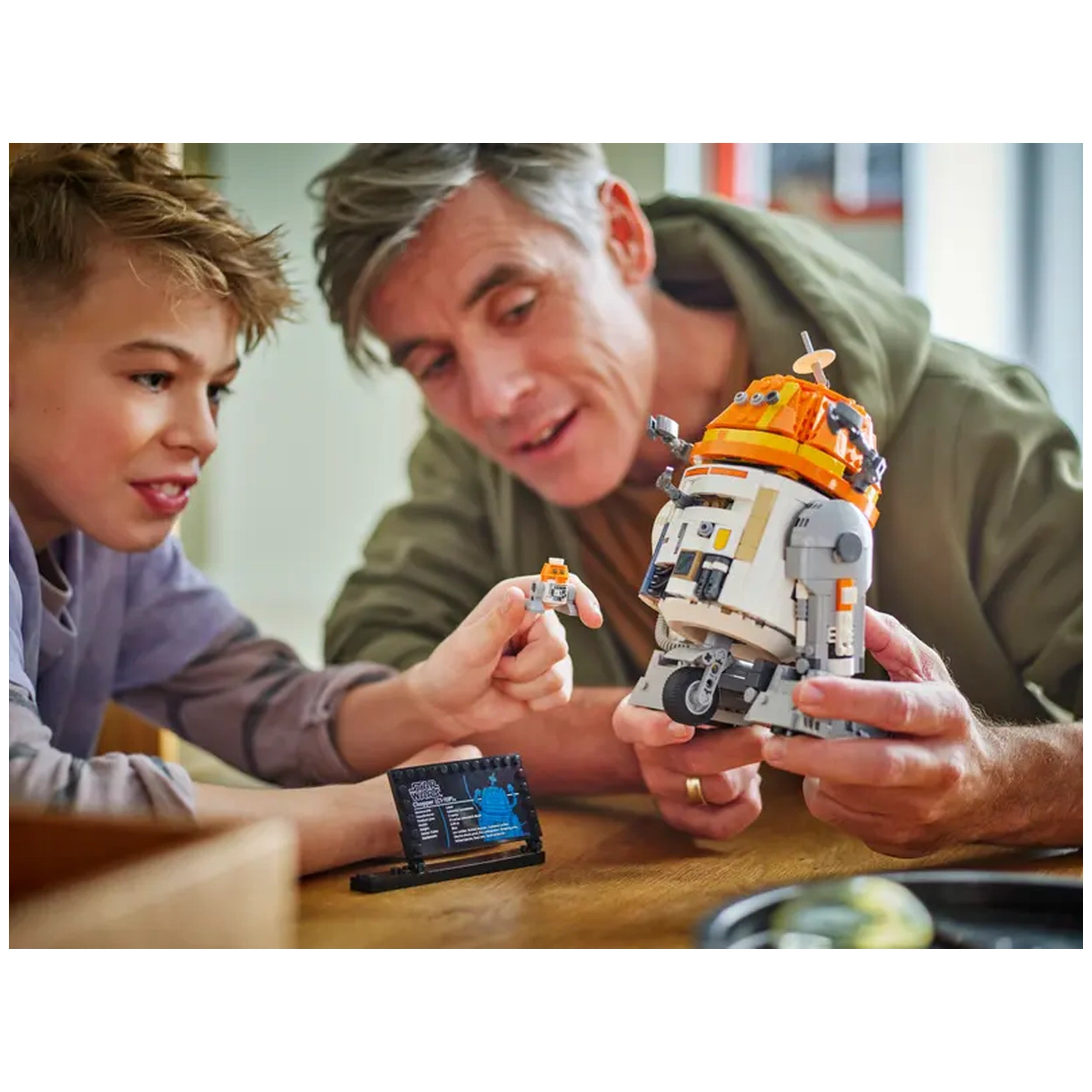 A young boy and a man joyfully interact over a LEGO Chopper (C1-10P)™ astromech droid, showcasing its intricate details. The boy points to a smaller LEGO figure, while the man holds the larger droid, both smiling. A build instruction booklet rests on the table, enhancing the playful atmosphere.