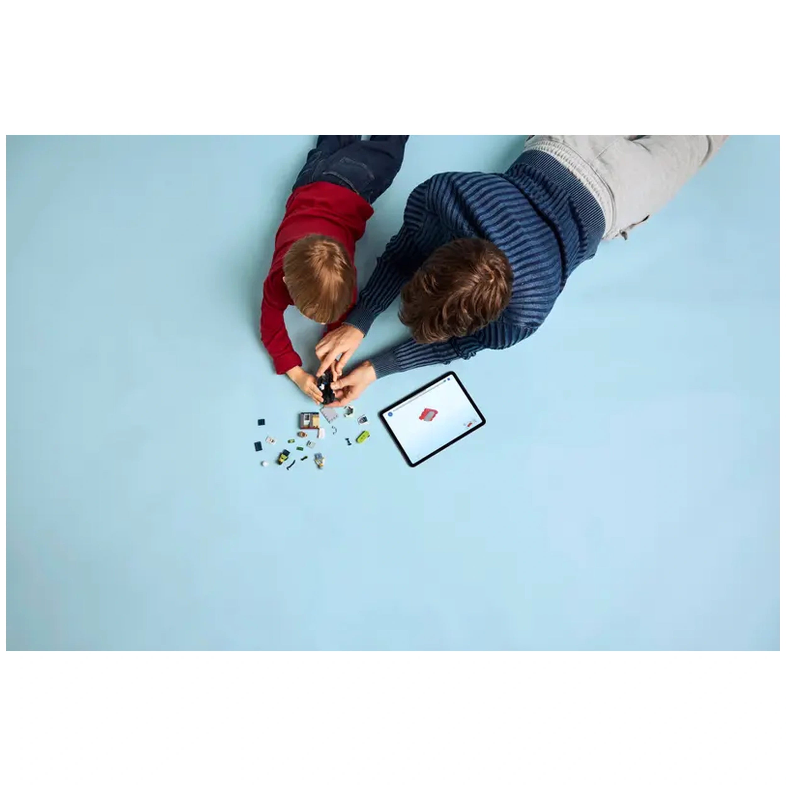 A child in a red shirt and an adult in a striped sweater work together on a LEGO set, surrounded by colorful bricks on a light blue floor, with a tablet nearby displaying building instructions.