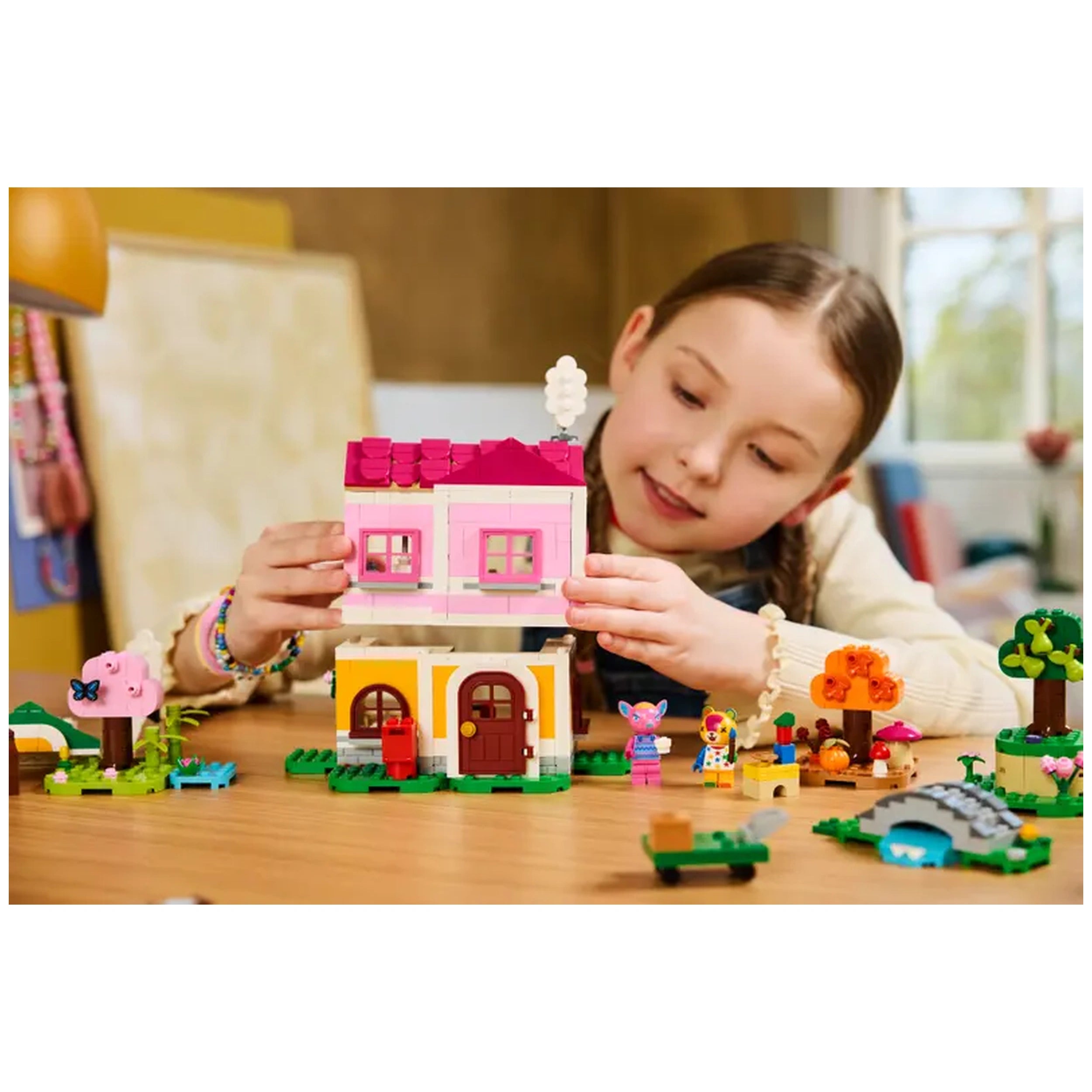 A young girl is focused on assembling a colorful LEGO house with pink and yellow details. Surrounding her are various accessories, including trees, a pink flower, and playful animal figures, on a wooden table. The scene showcases a vibrant, creative play environment inspired by Animal Crossing.