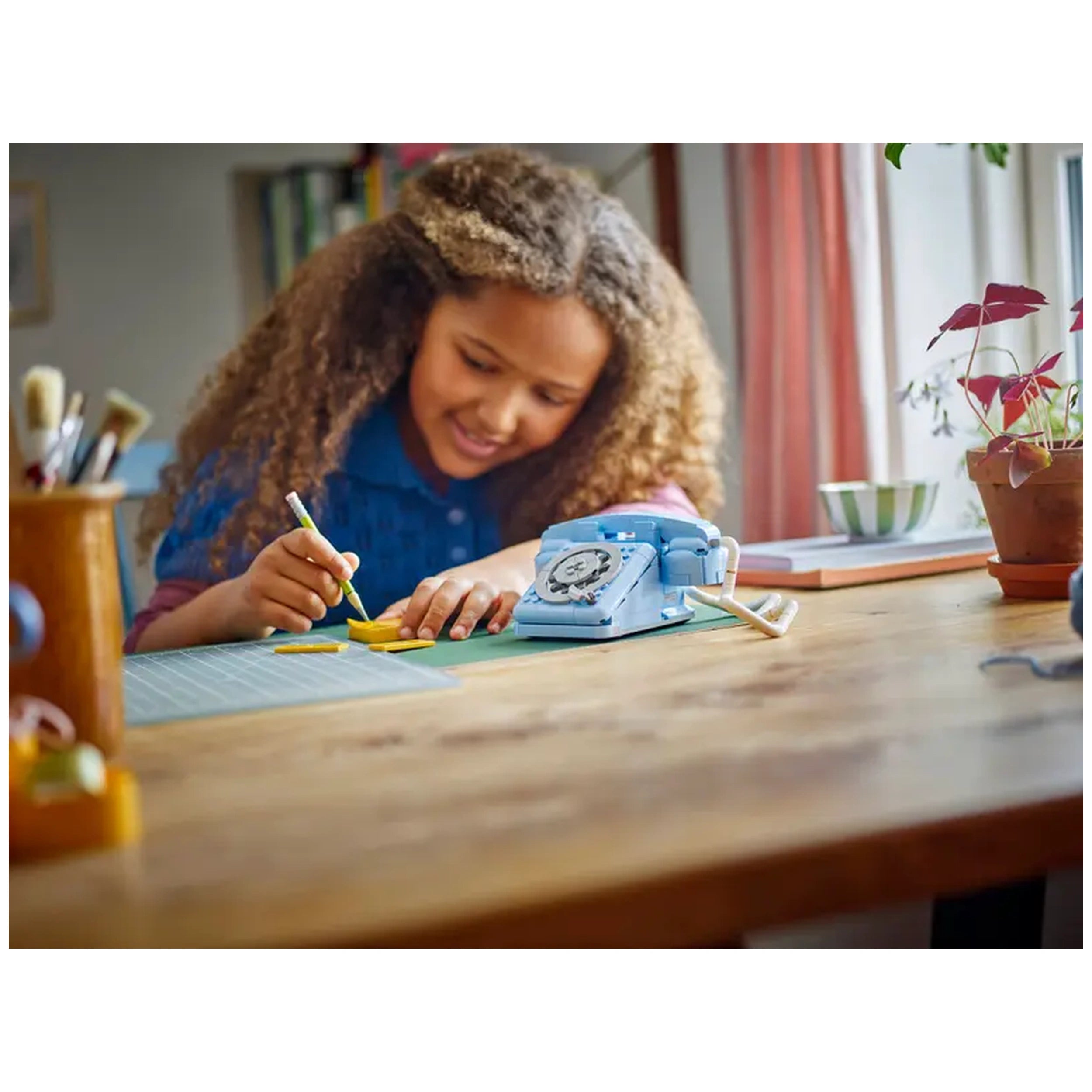 A girl with curly hair draws on yellow LEGO pieces next to a blue retro telephone on a wooden table, surrounded by art supplies.