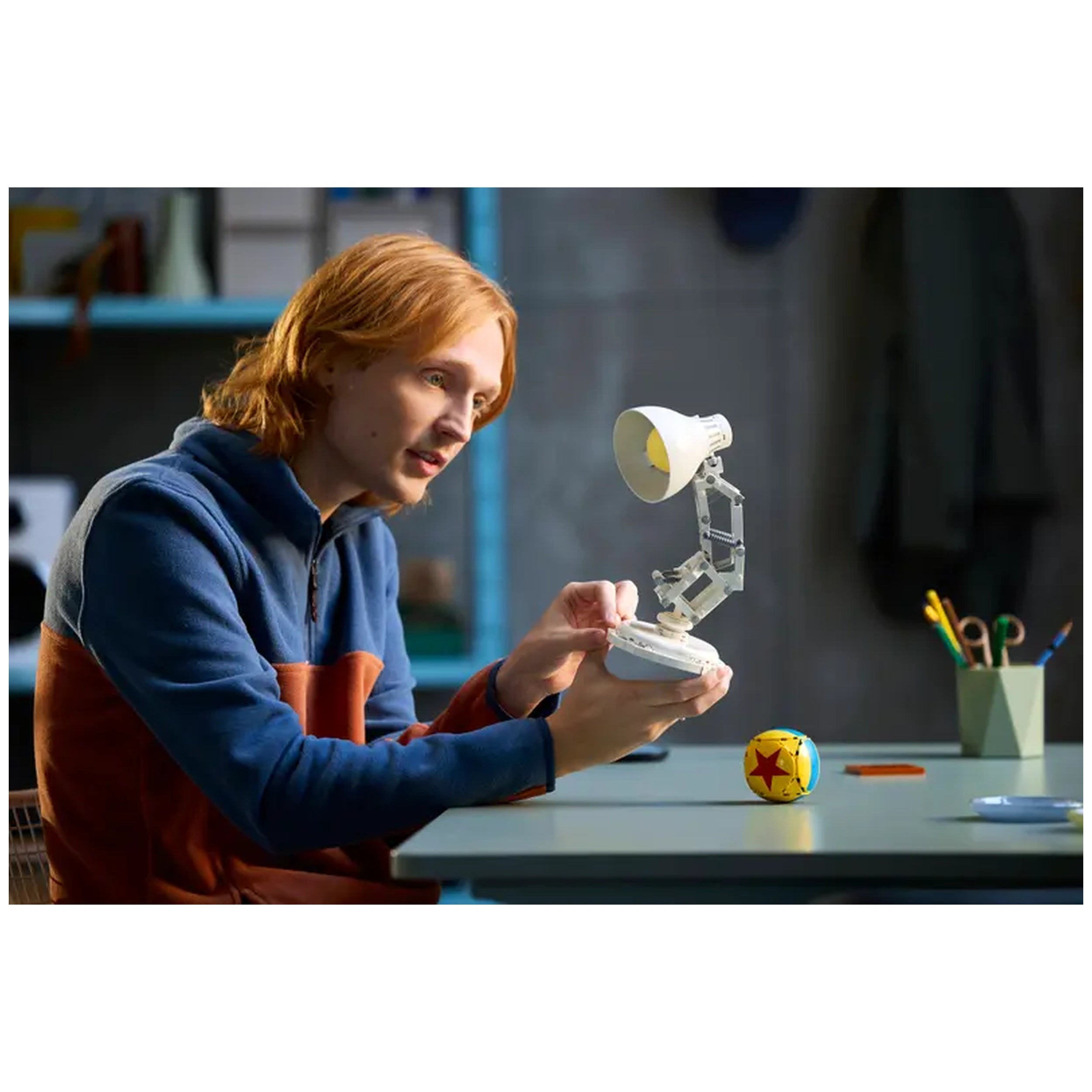 A young man with long, wavy red hair attentively examines the LEGO Disney Pixar Luxo Jr. model in a cozy workspace, featuring a colorful ball and stationery in the background.