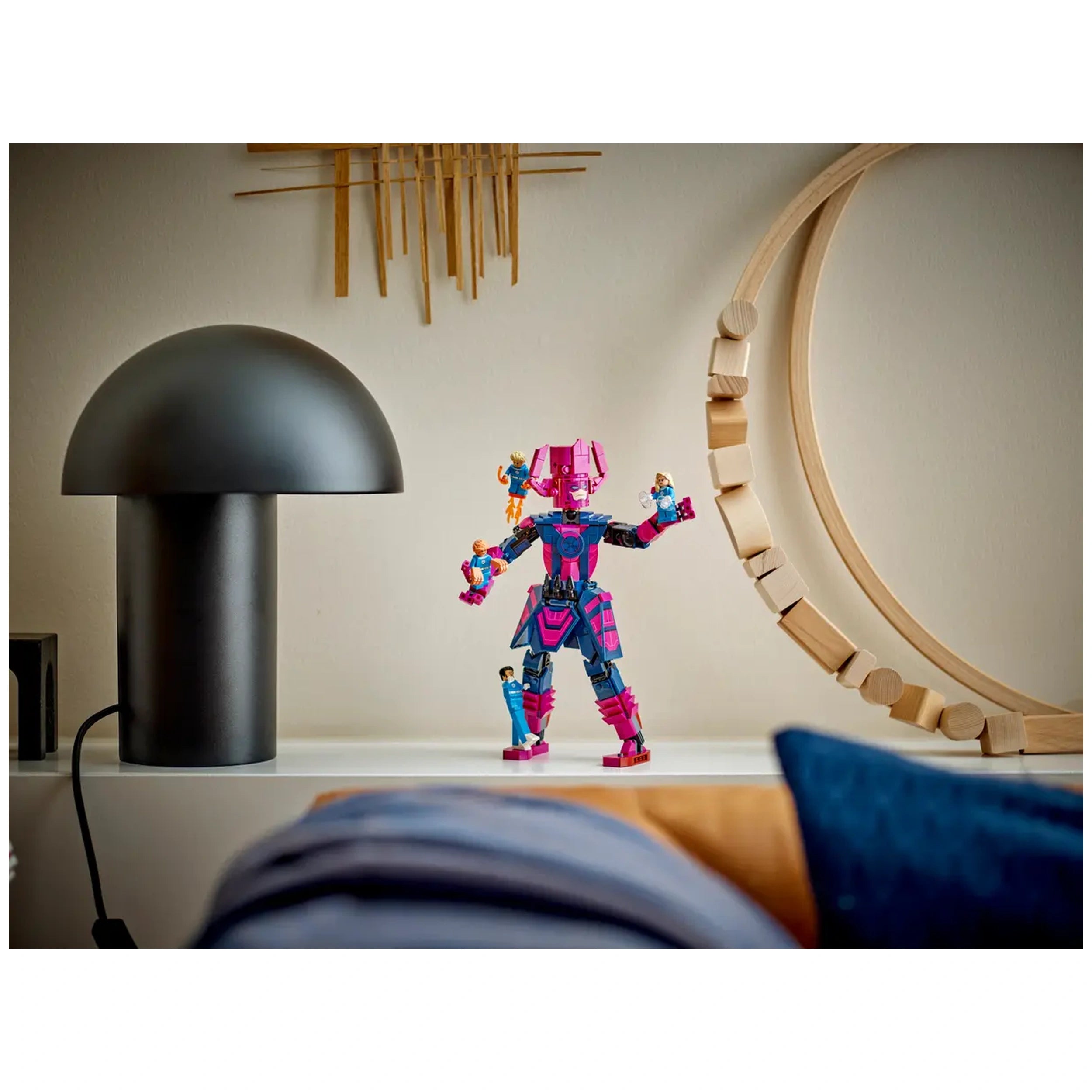 Superhero construction set featuring a brightly colored figure with a pink and blue design, holding small minifigures. A black lamp and wooden decor are in the background on a white shelf.
