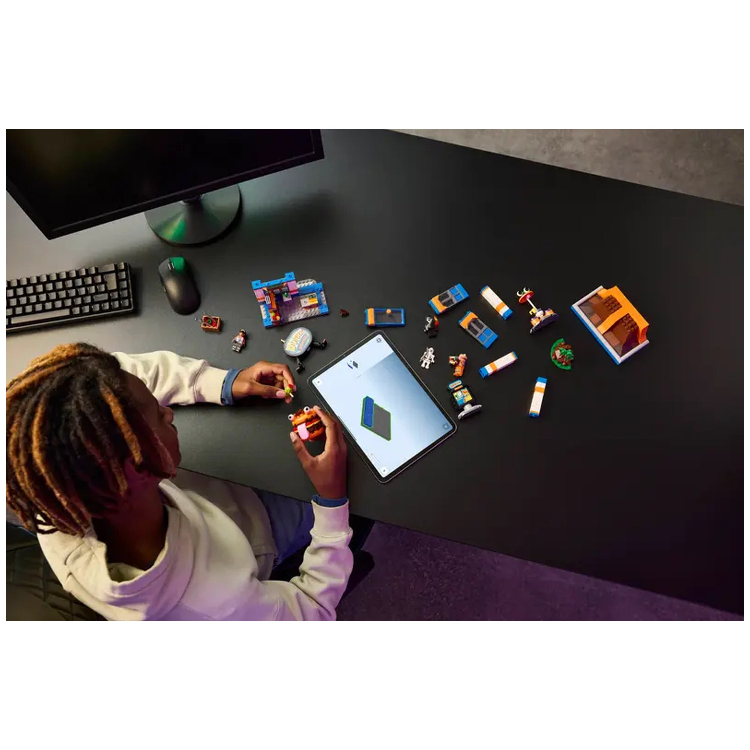 A child in a hoodie sits at a black table, constructing a LEGO Fortnite Durrr Burger Restaurant with various colorful pieces spread out. An open tablet displays building instructions, while a keyboard and mouse are in the background.