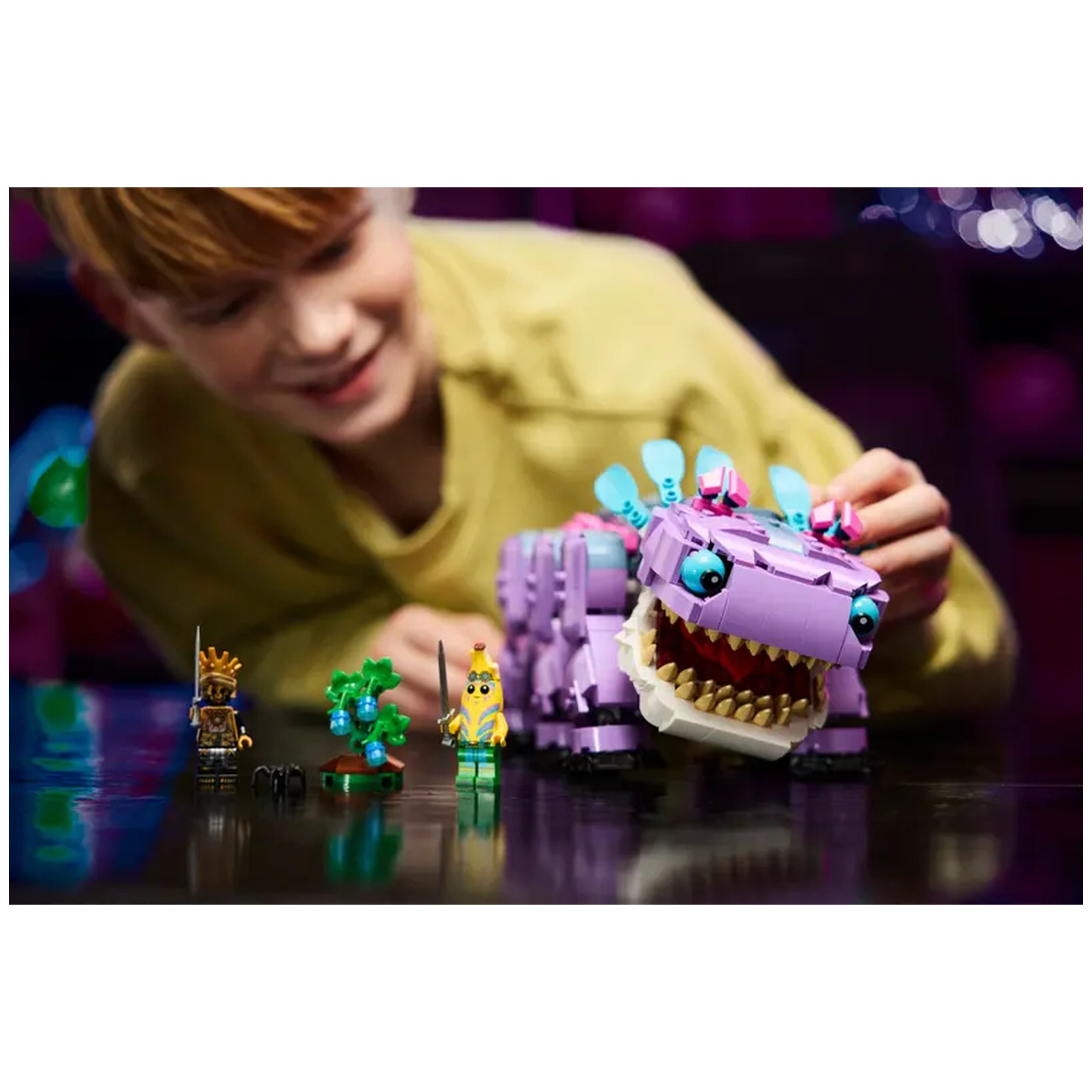 A child smiles while playing with the LEGO Fortnite Klombo construction set, featuring a large purple Klombo character and two minifigures, Island Adventure Peely and a green plant. The vibrant setting enhances the excitement of imaginative play.
