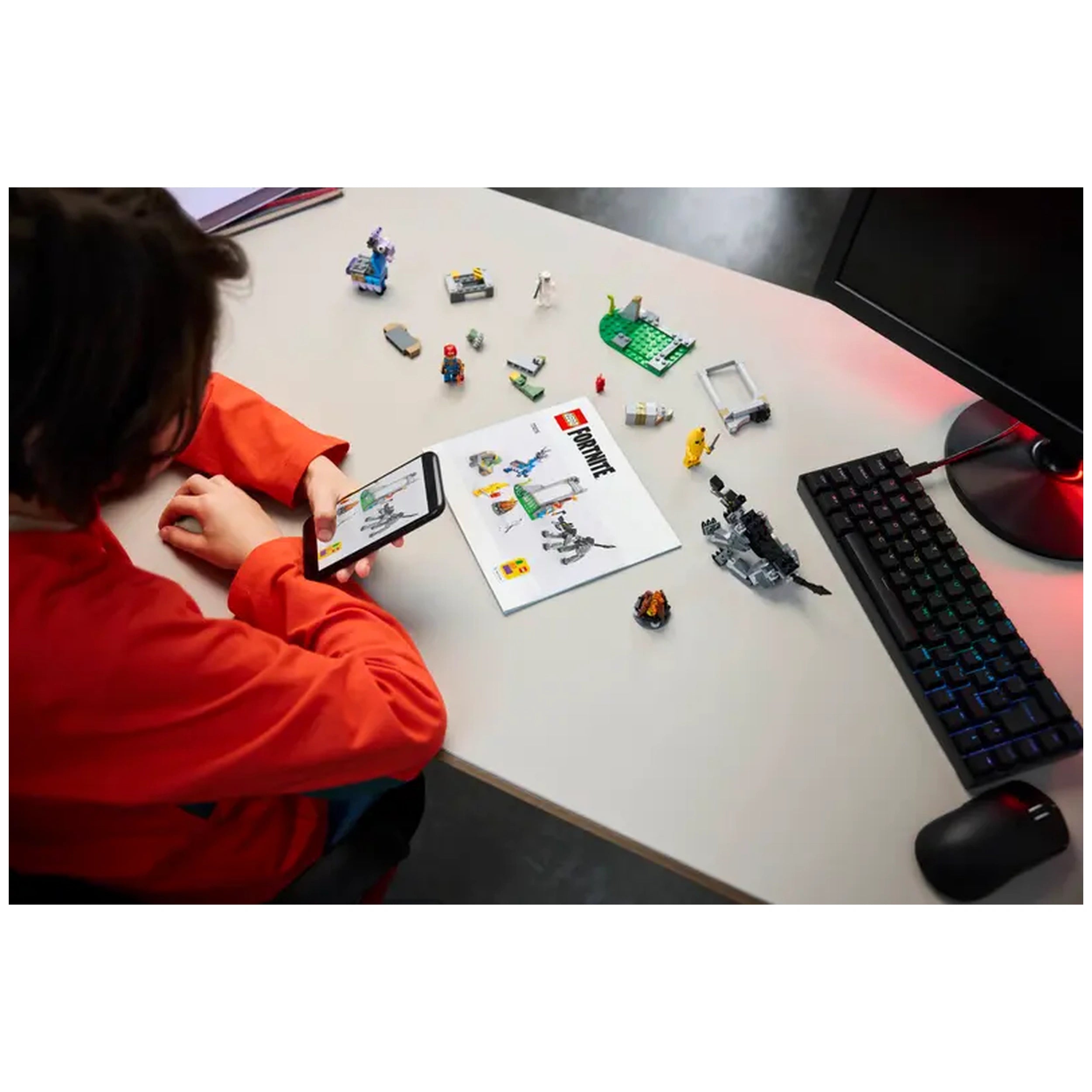 A child in a bright orange shirt sits at a table, looking at a smartphone while referencing a LEGO Fortnite instruction manual. Scattered around are various LEGO pieces, including figures and components from the Peely & Sparkplug’s Camp set. A keyboard and computer mouse sit on the edge of the table, adding to the playful atmosphere.