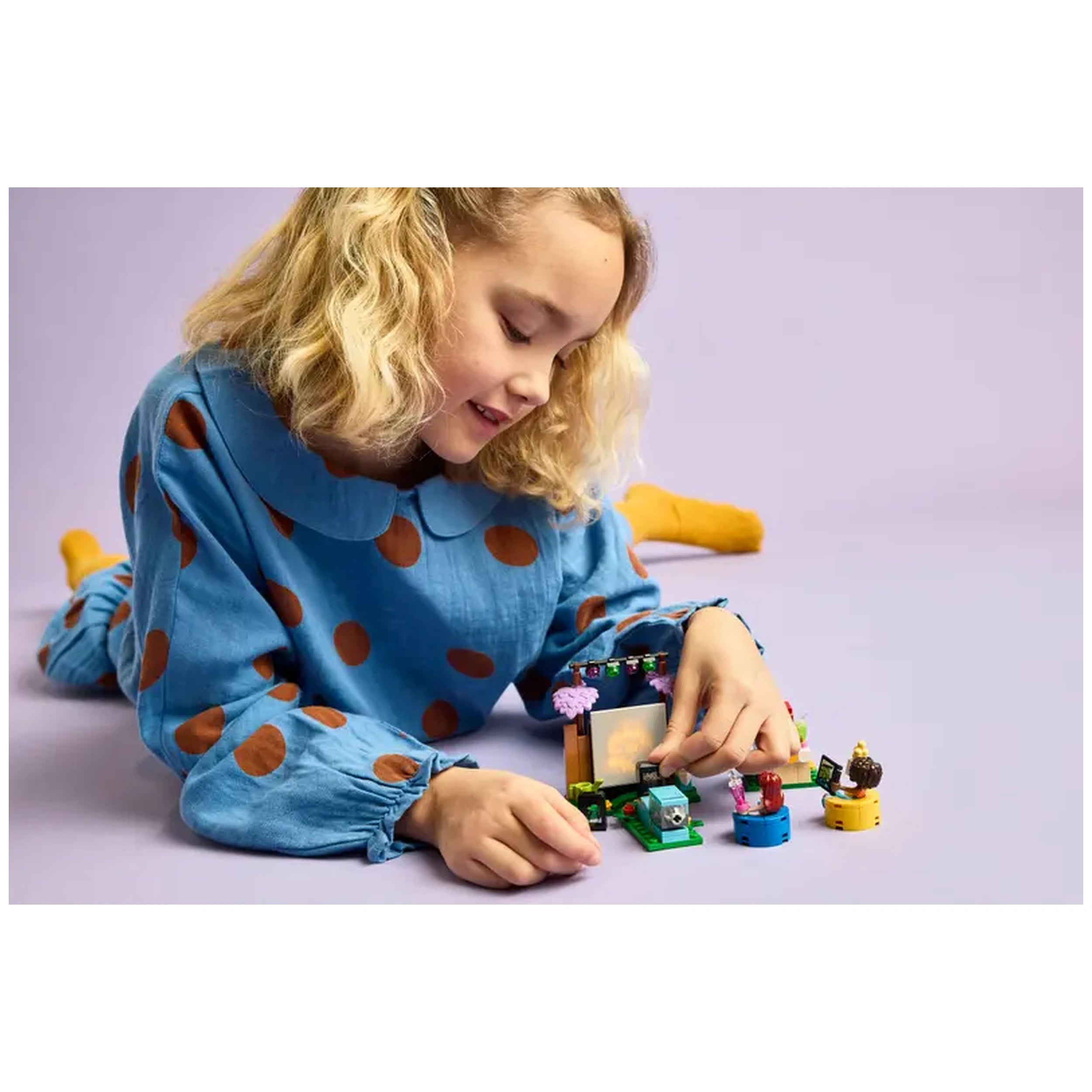 A young girl, wearing a blue polka-dot outfit, smiles as she plays with the LEGO Friends Friendship Movie Night set on a purple background. She is focused on placing movie slides in the small projector while colorful accessories surround her.