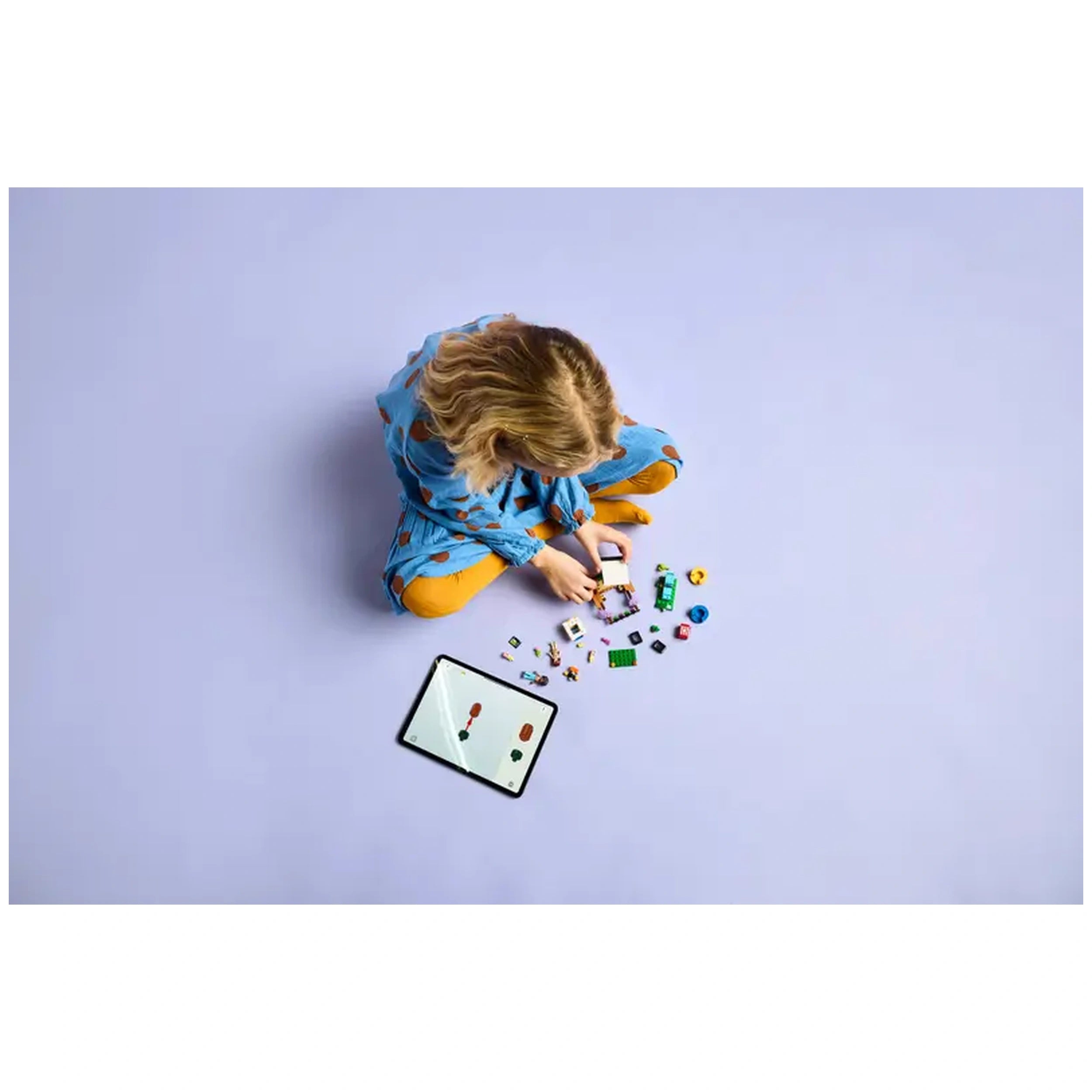 A child sits cross-legged on a lavender floor, engaged with LEGO pieces scattered around. An iPad displays colorful building instructions, while the child carefully assembles a toy.