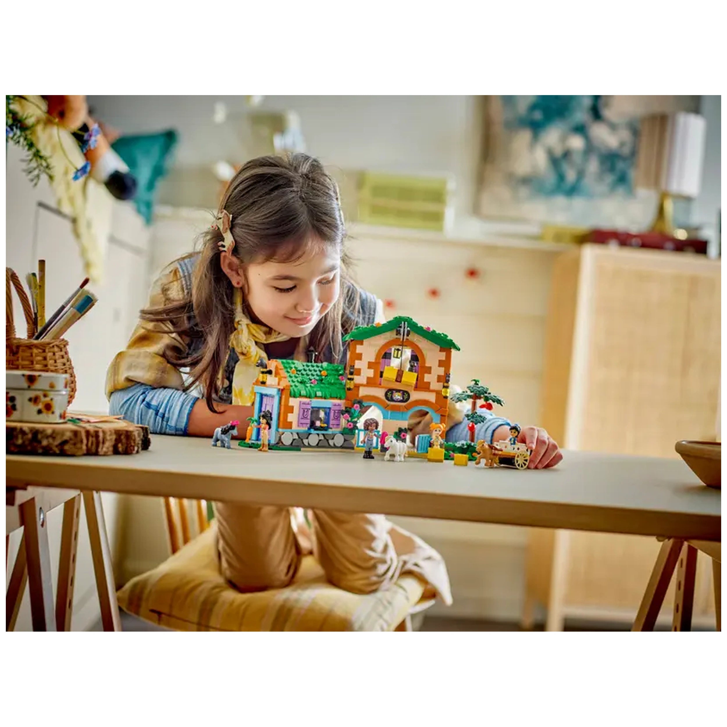 A girl with long brown hair leans forward on a table, engaging with the colorful LEGO® Friends Pony Ranch & Stable set, featuring stables, characters, and animals. The bright scene is set in a cozy room with warm lighting and rustic decor.